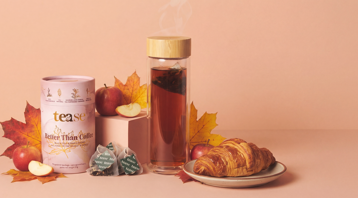 A styled tea product scene on a warm peach background, featuring a canister of 'tease Better than coffee' tea, several tea bags, a glass bottle of steeping tea with a bamboo lid, apple slices, fall leaves, and a croissant on a plate.